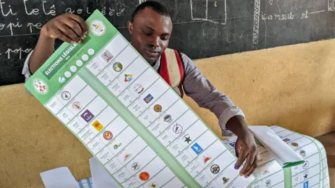 AFP/Getty Images Um funcionário eleitoral masculino prepara um boletim longo com pelo menos 28 candidatos a um eleitor em uma assembleia de voto em uma sala de aula na Escola Primária Pública de Hedzranawoe - um quadro -negro pode ser visto atrás dele - em Lomé em 29 de abril de 2024 durante as eleições legislativas do TOGO.