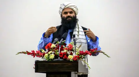 Sirajuddin Haqqani addresses the audience from a podium covwered in white and red flowers. 