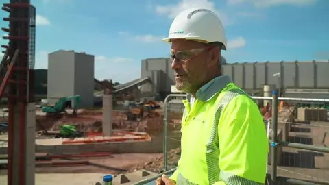 Jeff Court is wearing a high-vis top and hard hat. He stands in front of grey industrial buildings and orange soil which has been mined.