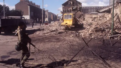 PA Media An Army soldier patrols a section of the Falls Road in Belfast. Beside him is a large bundle of barbed wire, and in the foreground, a yellow digger is digging up rubble left behind after a blast destroyed part of a building.