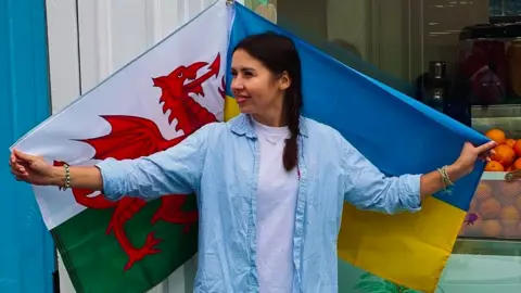 Maryna Korolova Maryna holding Wales and Ukraine flags. She is wearing a blue and white striped shirt with a white shirt underneath, and her long dark hair is in a plait. A box of oranges can be seen in the background.