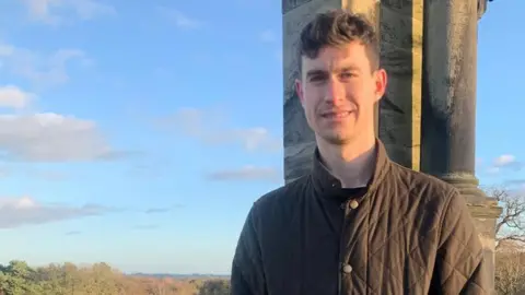 Chris Whittock, standing outside with countryside behind him and a blue sky. He is wearing a buttoned-up brown jacket.