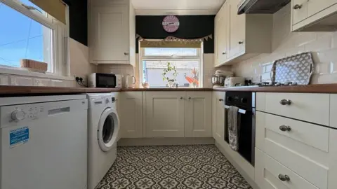 Rightmove photograph of cream cupboard cabinets in a small kitchen. A fridge and washing machine can be seen on the left. Drawers and a built in oven can be seen on the right. There are mosaic tiles on the floor and a window