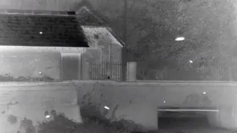 Ian Baker Thermal image of a small brick structure near a bridge, showing heat variations across the roof, walls, and surrounding vegetation, with cooler tones dominating the scene. White spots across the picture indicate where small bats are.