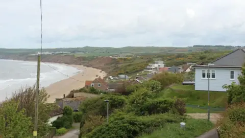 Anttoni James Numminen/LDRS A number of cliff-top homes close to an eroded green slope leading to a stretch of sand and sea