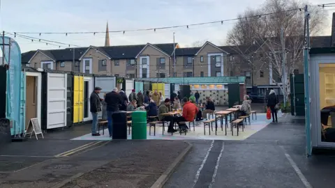 A number of people are sitting outside in a food and drink area, surrounded by brightly coloured shipping containers.