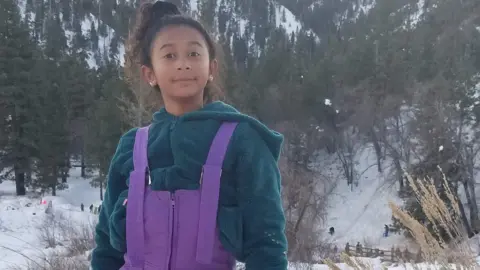 Celeste Rivas Hernandez is seen in a snow outfit. She is wearing a teal jacket and bright purple overalls 