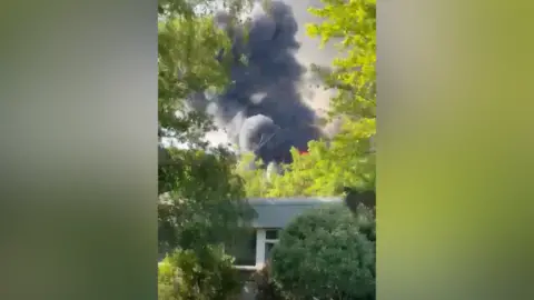 Paul King Smoke and flames rise above a white roofed building with trees around it.