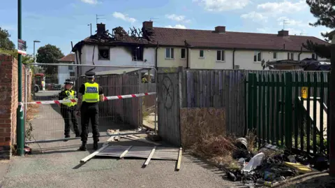 PA Police officers are standing at a metal fence sealing off an alleyway next to a badly burnt property. An 'x' in a circle can be seen next to a panel of fencing which is on the ground. The roof of the property is badly burnt.