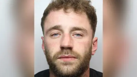 Police custody image of James Saxon with brown beard and hair wearing a black t-shirt.