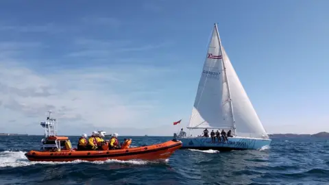 Ollie Burton/RNLI A small sailing ship sails past the Channel Island of Herm, flanked by an inflatable inshore lifeboat.