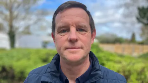 A man with dark hair looking at the camera he is wearing a navy coat