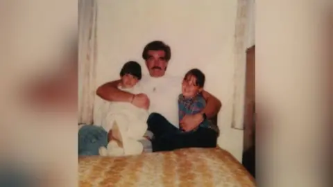 FAMILY HANDOUT An old photograph of Tony and his two daughters. They are sat on a bed and he has his arms around them