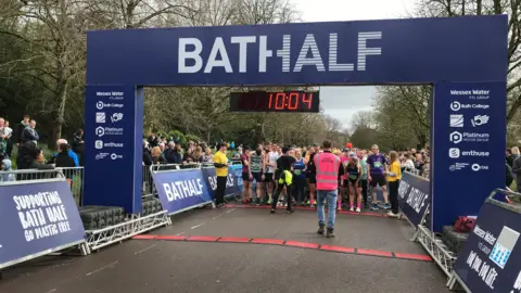 A queue of runners line up just behind an arch reading BATH HALF. A race marshal in a pink high-vis jacket stands in front. Spectators stand behind barriers to the side. 