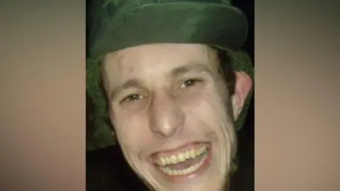 A man wearing a green cap is pictured grinning at the camera. He has brown eyes and some brown stubble around his cheeks and chin.