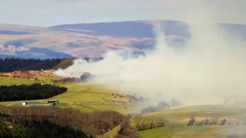 A huge pyre of sheep and cattle carcasses with suspected foot and mouth disease being burnt on the Epynt near Trecastle, 2001. Smoke billows out over surrounding hills and fields. 