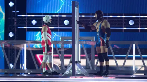 BBC/Hungry Bear Media Ltd/Graeme Hunter Mark Billings and Gladiator Giant are standing either side of a large perspex barrier on a raised platform.