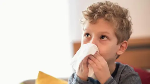 Getty Images Little boy with flu virus sitting on the sofa and wiping his nose with a napkin.