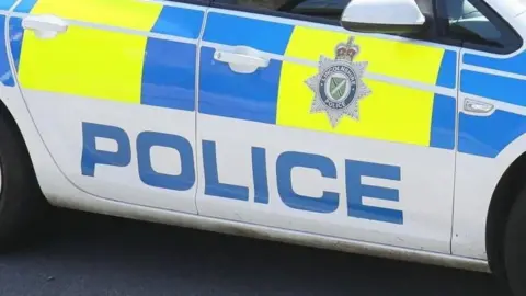 Side view of a marked Lincolnshire Police car it is white with yellow and blue blocks and the force's badge on the door