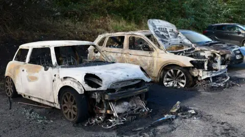 A wide view of three cars which have been destroyed by fire. Two of the cars are completely burnt out, the third on the far right is an Audi and is badly damaged but still recognisable