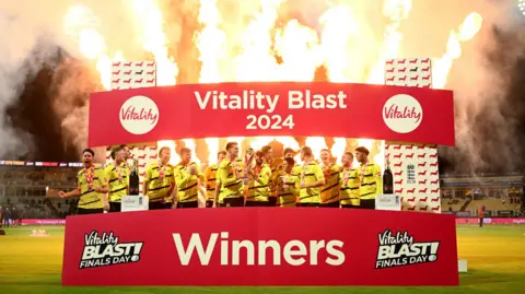 Getty Images Gloucestershire cricketers celebrate winning the T20 blast final against Somerset at Edgbaston. They are standing on a plinth with fireworks going off overhead, and a large sign with red background and white writing says "T20 blast champions"