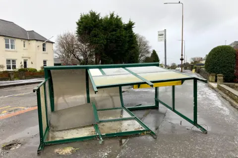 PA Media A smashed up green and glass bus stop lies on its side on a pavement. There are houses in the background.