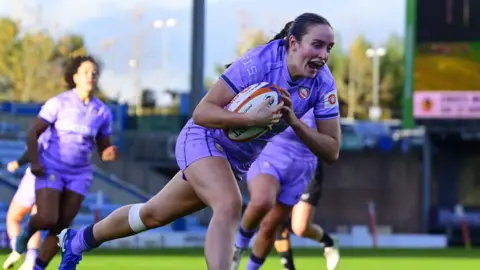 Nel Metcalfe bends towards the ground as she runs towards the try line with the ball clasped to her chest to score a try