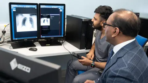Two men are looking at a screen on the left. The screen has pictures of chest x-rays. One man is wearing hospital scrubs while the other is wearing a grey suit, white shirt and blue tie. 