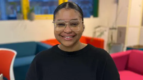 Anya in a black jumper. She is smiling and wears glasses.