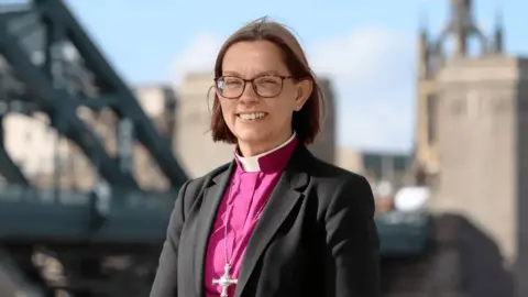 Diocese of Newcastle A portrait picture of Helen-Ann Hartley, bishop of Newcastle. She is smiling and has bobbed brunette hair, tortoise-shell glasses and studded earrings. She is wearing a purple clerical shirt and collar. a black suit jacket and crucifix around her neck. She is pictured outside with city buildings behind her and a blue sky.