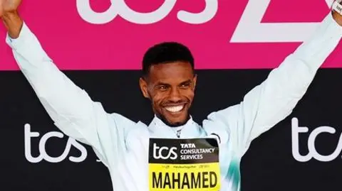 Mahamed Mahamed holding his arms aloft, smiling. He is wearing a white, long-sleeved top. He is standing in front of pink and black banners for the tcs London Marathon. He is wearing a yellow and black sign on his chest saying 'Mahamed'.
