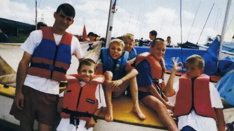 Anton Honchanka Anton sits on a boat with the Amoss family. They are all wearing life jackets and there are people stood behind them with the masts sticking in the air from other boats.