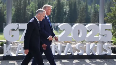 EPA Canadian Prime Minister Mark Carney and US President Donald Trump walk in front of a large 3D sign that says G7 2025 in white, stacked on top of the word Kananaskis. Behind the sign are tall green pine trees, and an expanse of green lawn.