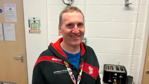 Matt Fisher, a community worker at the charity Up for Yorkshire, is looking straight into the camera. He is wearing a multi-coloured lanyard and a red-and-black tracksuit top