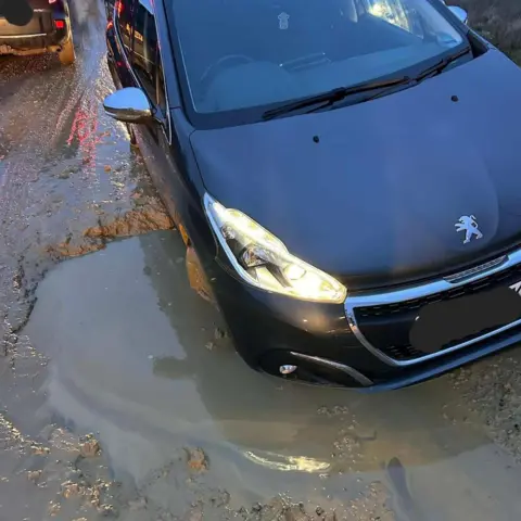 LDRS A black Peugeot car stuck in a large pothole filled with water on a road.