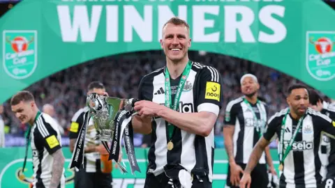 DANEHOUSH/Getty Images Dan Burn tersenyum ke kamera yang memegang trofi Piala Carabao di bawah lengkungan 'pemenang' hijau di pitch Wembley. Rekan satu timnya dapat dilihat di latar belakang.