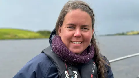 Headshot of Amy Burns smiling into the camera. She is wearing a life-jacket. In the background a body of water and beyond that a grassy hill.