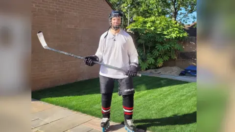 Will Chadwick Will Chadwick in skating ear and holding his hockey stick. He is standing in a garden with grass and trees behind him. 