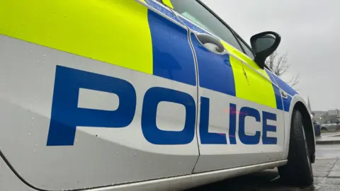 The image shows the side of a police car, close up. The vehicle has yellow and blue stickers on it. A large police logo is displayed down the side of the car.