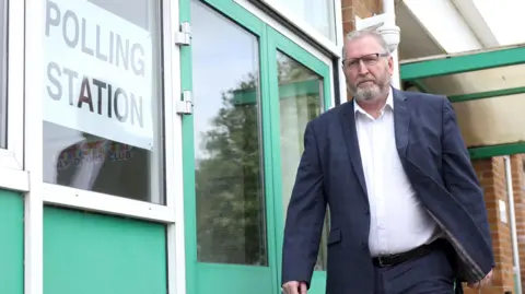 Pacemaker Man in blue suit and white shurt with beard and glasses walking by a polling station 