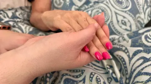 Two people are holding hands over a duvet cover. One hand is of an older woman with pink painted nails. The other hand is also a woman's.