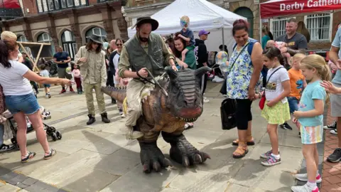 A man riding a plastic dinosaur while young children look on