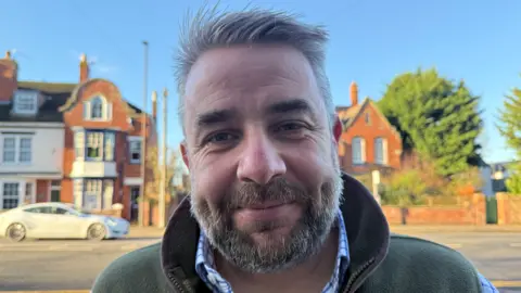 Richard Davies has grey hair and a grey and brown beard. He's wearing a green jacket and is standing in front of some houses.