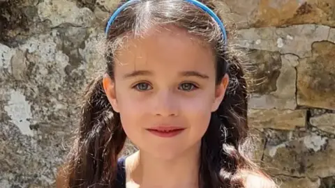 Lancashire Police Head and shoulders of a smiling Millie Gribble in front of a stone wall. She has long dark hair tied back in two bunches with a thin blue headband.