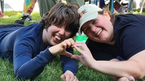 Two young people smile broadly at the camera as they lie on some grass and make a heart with their hands. 