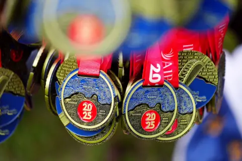 A collection of 2025 Great North Run medals. The medals are round with the medal divided into two sections, one coloured blue and one black with the outline of a map engraved on them and a round section coloured red with 2025 in them. The medals are on red ribbons