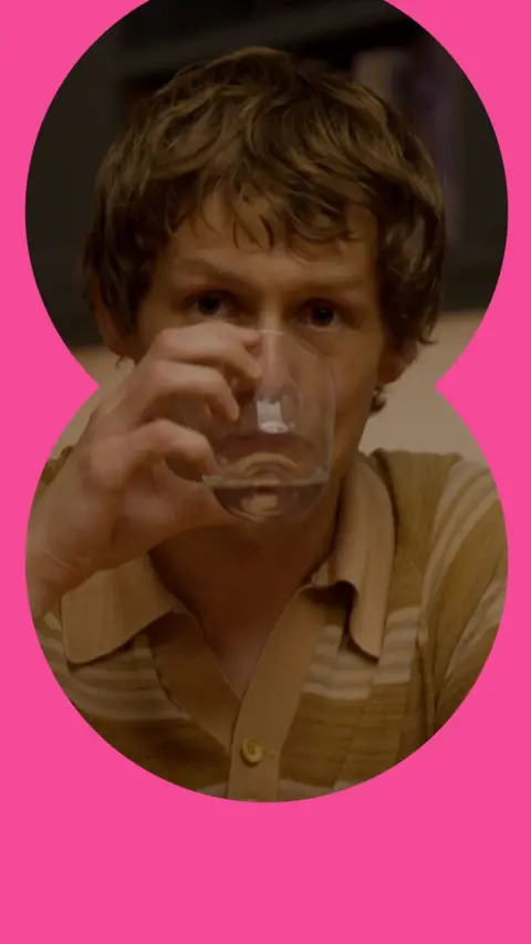 A man drinks from a glass at a dinner table. He has short brown hair and wears a brown patterned T-shirt