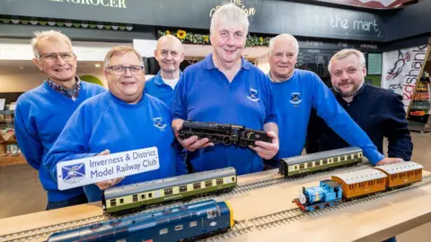 Paul Campbell A group of six men stand at a table with some sections of track and locomotives and carriages. Five of the men are wearing the same blue colour of tops with logos of Inverness and District Model Railway Club.