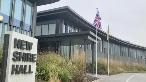 Brian Farmer/BBC A council building with a large square stone sign outside saying "New Shire Hall" in dark grey lettering against a beige background. Two flagpoles outside fly the British flag and a yellow and blue one. There are decorative grasses outside the building which is modern with glass and vertical shading.