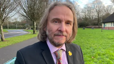 Mr Cox, who has shoulder-length hair and a short greying beard, smiles sadly at the camera. He is sat on a bench in a park, with a road behind him. He is wearing a black suit with a pink shirt and a red paisley tie, and a yellow duck pin attached to his lapel. 